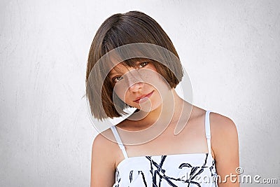 Pretty little girl with freckles, dark eyes and short hairstyle wearing summer white dress posing against white background having Stock Photo