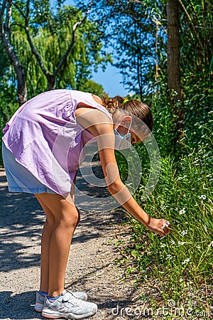 Pretty little girl with a mask to protect herself from the coronavirus picking a flower on a path surrounded by nature Stock Photo