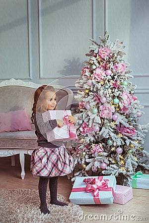 Pretty little girl stands with a gift box at hands near Christmas tree and looks to side Stock Photo