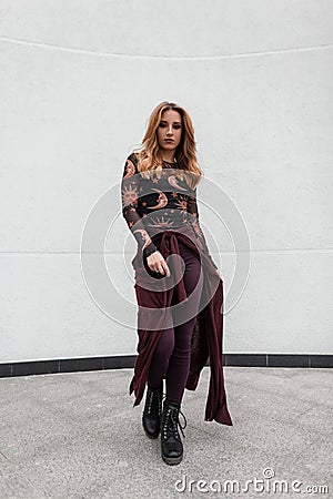 Pretty luxurious young woman with purple cape in a vintage patterned blouse in stylish pants in boots posing near a white wall Stock Photo