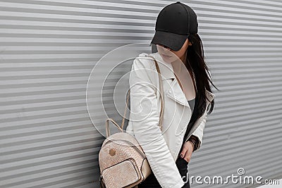 Pretty modern young woman in a fashionable black baseball cap in a vintage white leather jacket in jeans with a gold backpack Stock Photo