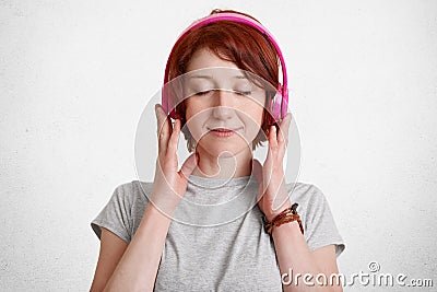 Pretty relaxed pleased ginger woman with short hair, closes eyes with pleasure while listens her favourite music with headphones, Stock Photo