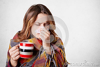 Pretty sick woman has runnning nose, rubs nose with handkerchief, drinks hot beverage, wrapped in warm blanket, has high temperatu Stock Photo