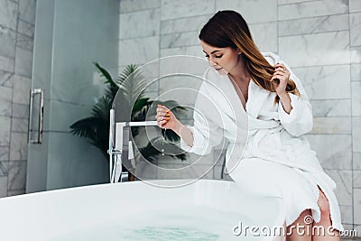 Pretty slim woman wearing bathrobe sitting on edge of bathtub filling up with water Stock Photo