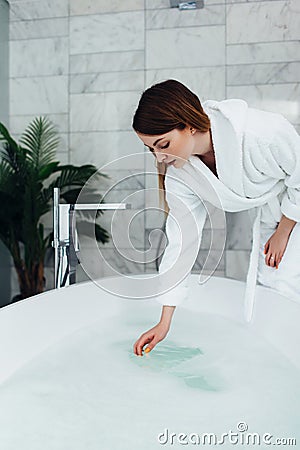 Pretty slim woman wearing bathrobe sitting on edge of bathtub filling up with water Stock Photo