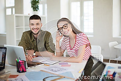Pretty smiling girl in striped t-shirt and eyeglasses happily closing eyes while working with colleague. Group of cool Stock Photo
