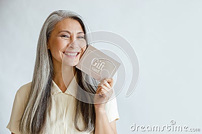 Pretty smiling mature Asian lady holds gift card on light grey background in studio Stock Photo