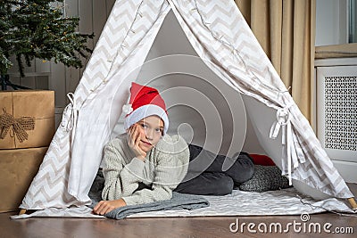 A pretty teenage boy in a Santa hat lying in a children& x27;s tent in a room waiting impatiently for Santa. Stock Photo