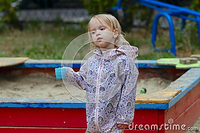 Pretty toddler girl wearing rain jacket playing with blue cup near the sandbox on the playground Stock Photo