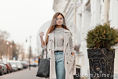 Pretty trendy young woman in a stylish trench coat in a vintage knitted sweater in blue jeans with a leather bag enjoys a walk Stock Photo
