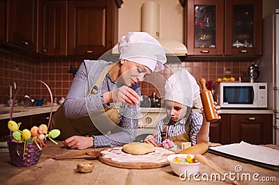 Pretty woman in chef& x27;s hat and beige apron, flouring dough, preparing Easter cake with her daughter in the home Stock Photo