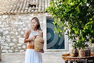 Pretty woman in ethnic Mediterranean folk traditional costume holding a rattan olive oil jug. Hospitality and ethnic tourism conce Stock Photo