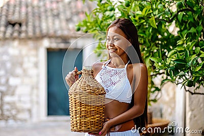 Pretty woman in ethnic Mediterranean folk traditional costume holding a rattan olive oil jug. Hospitality and ethnic tourism conce Stock Photo