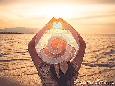 Pretty woman holding hands in heart shape framing setting during sunset on ocean beach Stock Photo