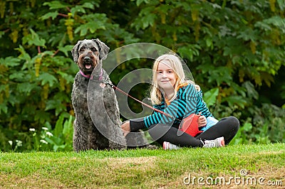 Pretty young blonde girl making eye contact while sitting on grass with well behaved black labradoodle dog on a leash Stock Photo
