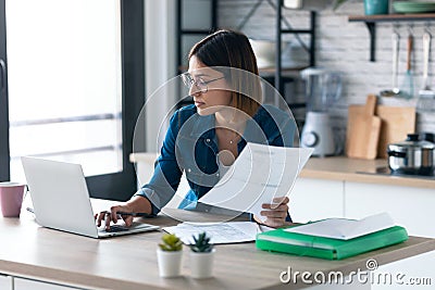 Pretty young business woman working with computer while consulting some invoices and documents in the kitchen at home Stock Photo