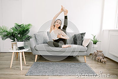 Pretty young Caucasian yoga fit woman sitting on gray sofa and posing to camera, stretching leg up, smiling and holding Stock Photo