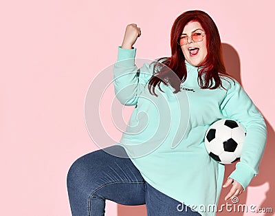 Pretty young overweight woman in jeans, blue hoodie and sunglasses standing with ball holding hand and knee up Stock Photo