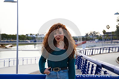 Pretty young red-haired woman is leaning on a railing in a park. She is posing for the photo smiling. Fashion and beauty concept Stock Photo