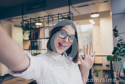 Pretty young smiling girl in glasses taking a selfie working in light room workplace workstation grimacing saying hello hi to her Stock Photo