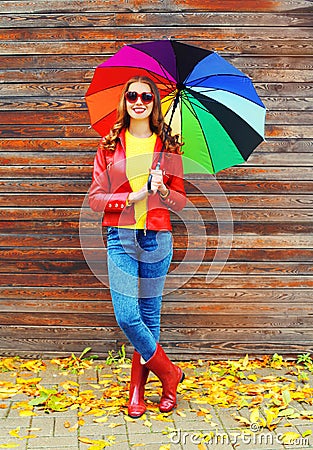 Pretty young smiling woman with colorful umbrella wearing a red leather jacket and rubber boots in autumn over wooden background Stock Photo