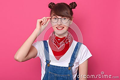 Pretty young woman with pleasant appearance, wearing white casual t shirt and overalls, having two hair buns, looks directly at Stock Photo