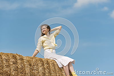 Pretty young woman sitting on haystack, countryside on blue background. Portrait of girl on high haystack Stock Photo