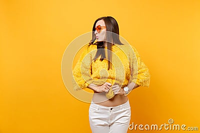 Pretty young woman in white pants, heart orange glasses holding taking off fur sweater looking aside isolated on bright Stock Photo