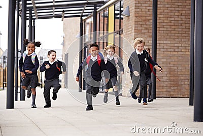 Primary school kids, wearing school uniforms and backpacks, running on a walkway outside their school building, front view Stock Photo