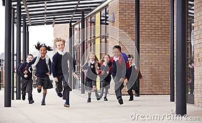 Primary school kids, wearing school uniforms and backpacks, running on a walkway outside their school building, front view Stock Photo
