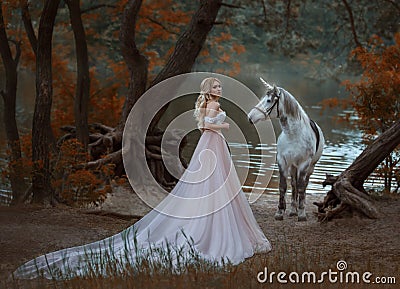 The princess met a unicorn in the forest. The blonde girl with a gentle make-up, is dressed in a long vintage dress with Stock Photo