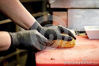 The process of making a burger in a restaurant kitchen. The chef in black gloves cut the bun. Stock Photo