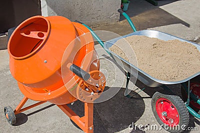 Process pouring the foundation. Orange concrete cement mixer machine is at construction site with wheelbarrow, tools, sand and Stock Photo