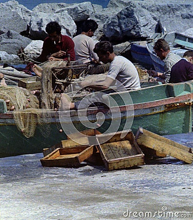 PROCIDA, ITALY, 1979 - Some fishermen repair their nets with skill and patience and dry some fish boxes in the Procida sun Editorial Stock Photo