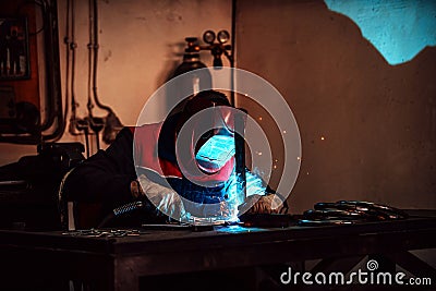 Profesional welder in protective uniform and mask welding metal pipe on the industrial table with other workers behind Stock Photo