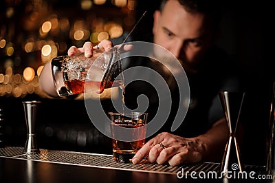Professional bartender pouring an alcohol from the measuring cup through the strainer to the glass with one big ice cube Stock Photo