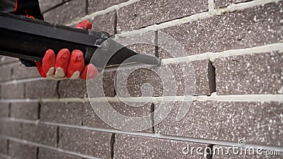 Professional builder filling fugue with grout gun near brick wall, closeup. Tiles installation process. Grouting with a Stock Photo