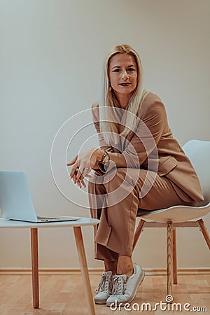 A professional businesswoman sits on a chair, surrounded by a serene beige background, diligently working on her laptop Stock Photo