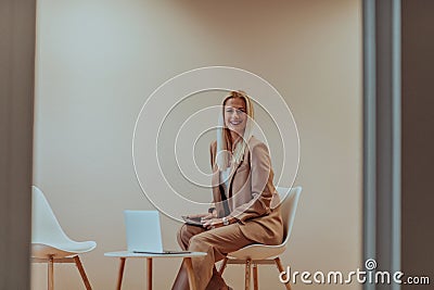 A professional businesswoman sits on a chair, surrounded by a serene beige background, diligently working on her laptop Stock Photo