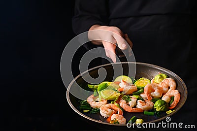 Professional chef cook on a dark background prepares seafood, shrimp with fire and vegetables. Roasting and cooking veggie food. Stock Photo