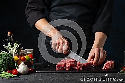 The professional chef cutting with knife fresh beef meat on the black board on dark blue background. Backstage of preparing rib- Stock Photo