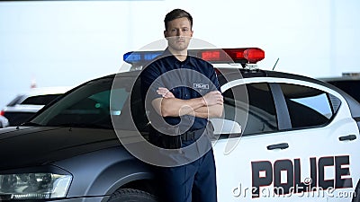 Professional policeman in uniform with crossed hands leaning on patrol car, duty Stock Photo