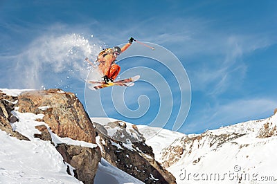 A professional skier makes a jump-drop from a high cliff against a blue sky leaving a trail of snow powder in the Stock Photo