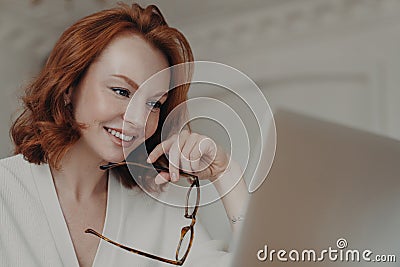 Professional smiling ginger female web designer uses application on laptop computer for creating project work, makes online Stock Photo