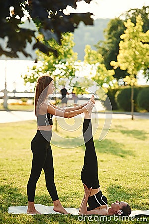 Professional trainer is helping. Two women in sport clothes are doing exercises outdoors on the field Stock Photo