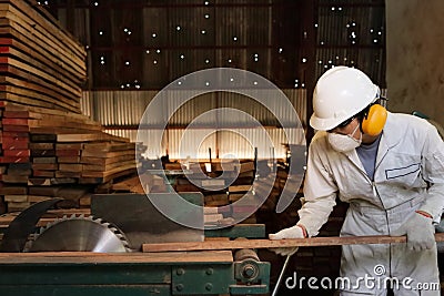 Professional young worker in white uniform and safety equipment cutting a piece of wood on table saw machine in carpentry factory. Stock Photo