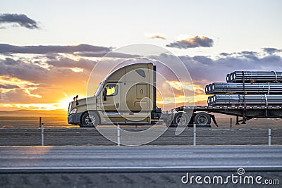 Profile of big rig beige semi truck transporting cargo on flat bed semi trailer driving on the one way highway road at sunset time Stock Photo