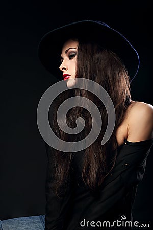 Profile of expressive female model posing in black shirt and elegant hat with red bright lipstick on dark shadow background. Stock Photo