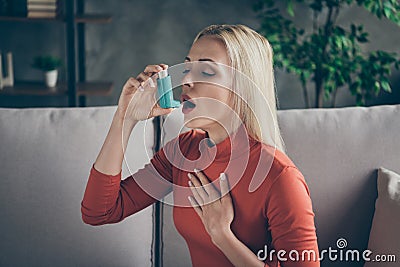 Profile photo of unhealthy pretty blond lady suffering allergy holding inhaler breathing in feel better sitting couch Stock Photo