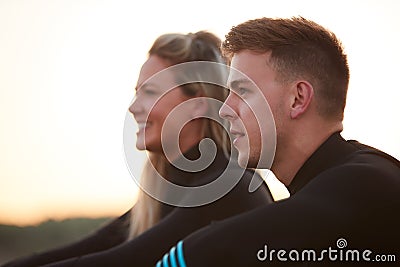 Profile View Of Couple Wearing Wetsuits On Surfing Staycation Sitting On Beach Looking Out To Sea Stock Photo